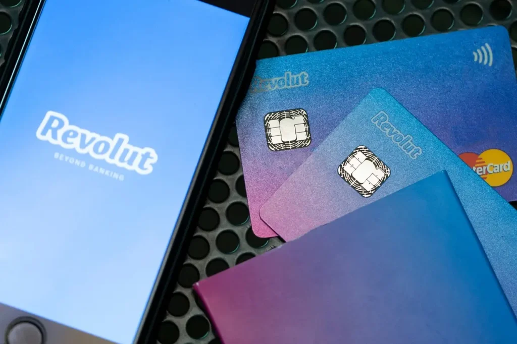 Smartphone mit Revolut App auf dem Bildschirm und rechts daneben Revolut Karten